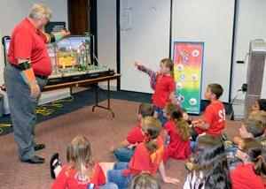 一名身穿红色衬衫的男子用桌面显示器向满屋子的小孩演示电气安全, 也穿着红衬衫.